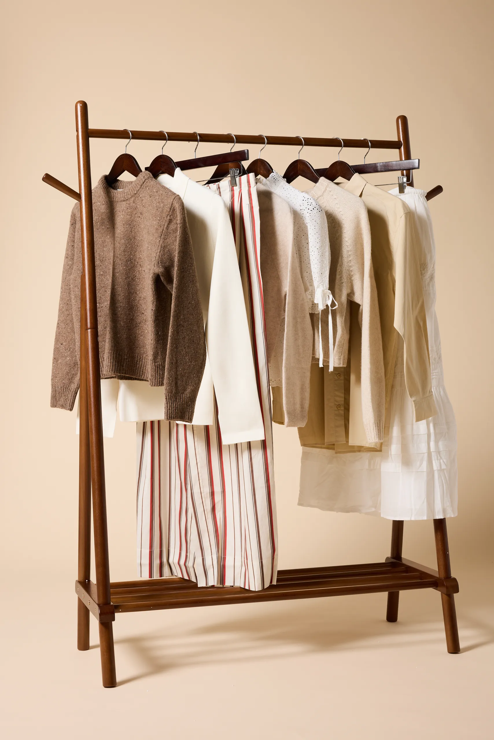 Wooden garment rack of neutral knitwear, shirts and striped trousers against a peach backdrop
